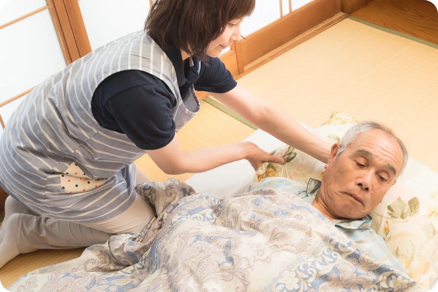畳の上の布団に寝ている高齢の男性の体の位置を変える看護師の写真